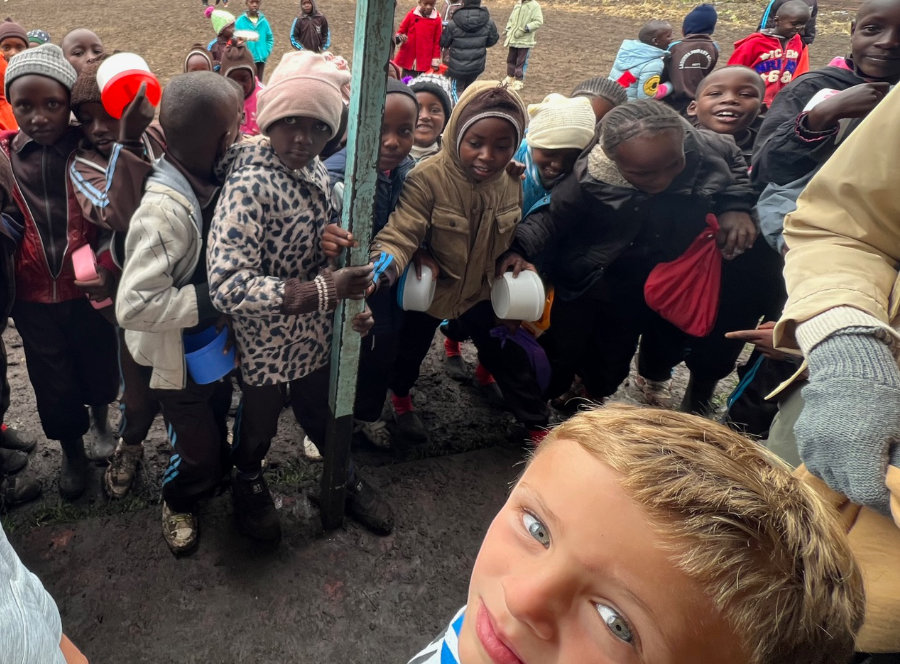 A group of children, some holding bowls and cups, gather outside. One child in the foreground looks up at the camera. The ground is muddy, and the children wear various jackets and hats.